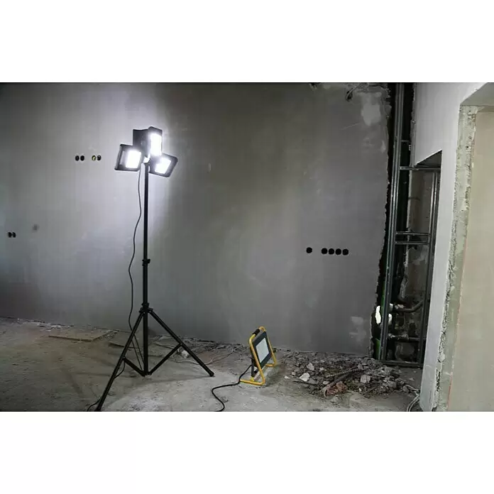 Profi Depot Proyector LED 2 cabezales - 50 W, 8.000 lm, 6.000 K - 7.000 K, L x An x Al: 1,14 x 1,32 x 2,25 m, IP54 - Foco de trabajo LED triple brillante sobre trípode negro iluminando una habitación sin terminar, con un foco de trabajo amarillo más pequeño en el suelo.