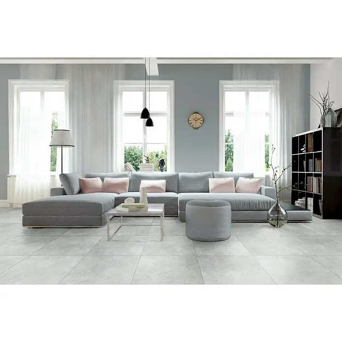 Feinsteinzeugfliese Beton - 60 x 60 cm, Grau, Matt - Wohnzimmer mit grauer Eckcouch, rosa Kissen, Couchtisch und Bücherregal. Helle Fenster und Lampen sorgen für eine freundliche Atmosphäre.