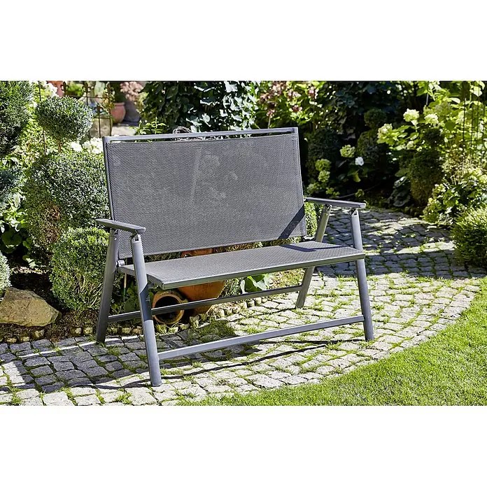 Sunfun Maja Banco de jardín 2 plazas - An x Pr x Al: 119 x 67 x 96 cm, Textilene, Antracita, Abatible - Un banco de jardín gris con diseño de malla se encuentra en un camino pavimentado rodeado de exuberante vegetación, ofreciendo una opción de asiento exterior elegante.