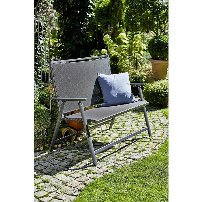 Sunfun Maja Banco de jardín 2 plazas - An x Pr x Al: 119 x 67 x 96 cm, Textilene, Antracita, Abatible - Un banco de jardín gris con un cojín azul se encuentra en un camino de piedra en medio de un jardín verde, invitando a la relajación. Mobiliario de exterior perfecto.