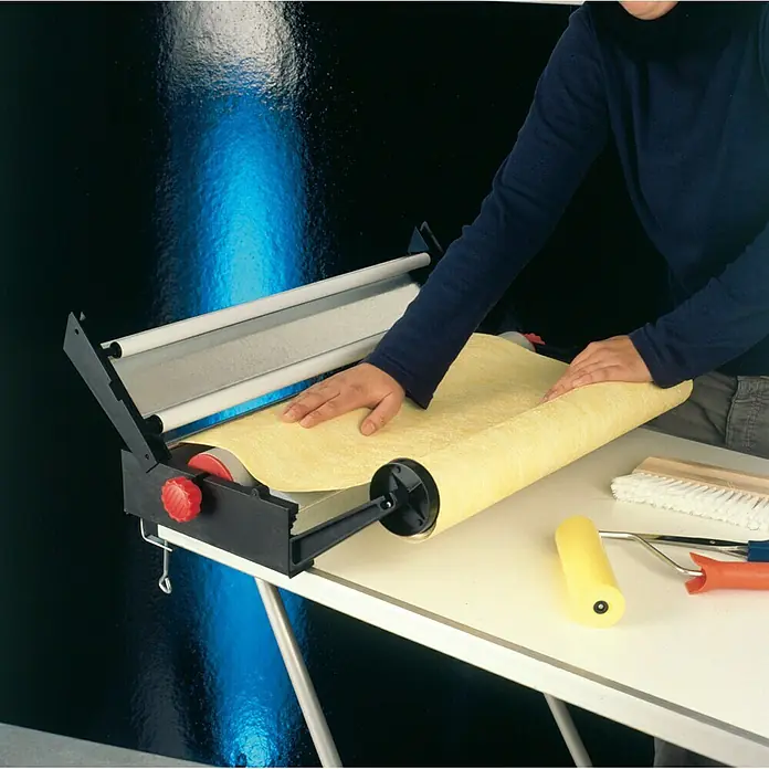 Decora Tapetenkleistergerät - Geeignet für: Euro Rollen 53 cm - Nahaufnahme eines Tapeziertisches mit einem Abroller, der Tapeten verarbeitet. Werkzeuge liegen bereit für die Anbringung.