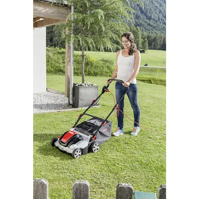 AL-KO Elektro-Vertikutierer Combi Care 36.8 E Plus Comfort - 1.400 W, Arbeitsbreite: 36 cm - Eine Frau mäht den Rasen mit einem AL-KO Rasenmäher in einem üppigen Garten, während ein langes orangefarbenes Kabel an den Mäher angeschlossen ist.