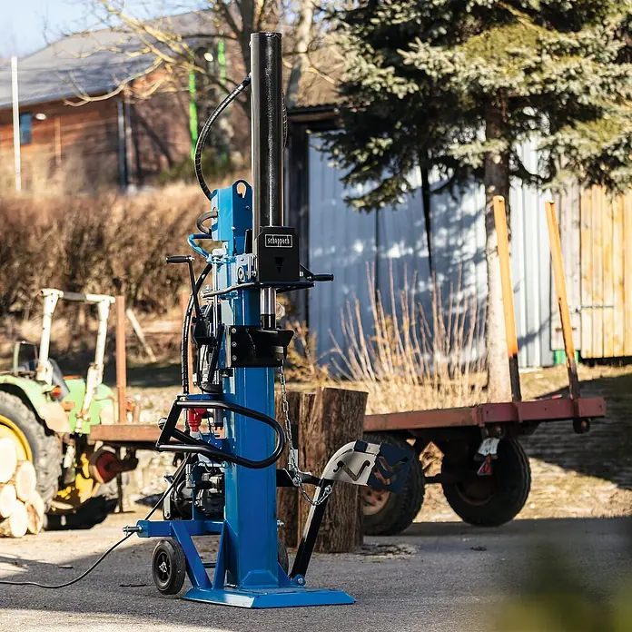 Scheppach Holzspalter HL3000GM - 30 t, Max. Spaltgutlänge: 110 cm - Eine blaue Scheppach Holzspalter Maschine steht im Freien mit einem Traktor und einem Anhänger im Hintergrund.