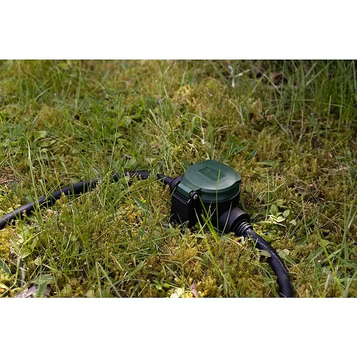 REV Verlängerungskabel - 10 m, Grün / Schwarz, IP44, H07RN-F - Ein grüner, wasserdichter Stromanschluss im Garten, umgeben von Gras und Moos, bietet eine sichere Stromversorgung im Freien.
