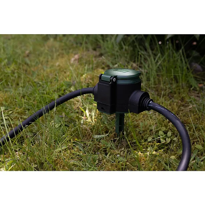 REV Verlängerungskabel - 10 m, Grün / Schwarz, IP44, H07RN-F - Eine im Gras installierte, wasserdichte Stromverteilerbox mit dunkelgrünem Deckel und schwarzen Kabeln für den Außenbereich.