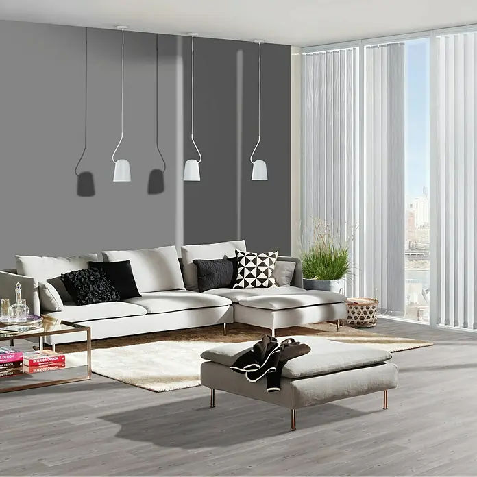 b!design Vinylbodenmuster Rigid SPC Swiss Pinie - 281 x 161 x 3,8 mm, Landhausdiele - Ein modernes Wohnzimmer mit einem grauen Sofa, einem passenden Hocker, stilvollen Pendelleuchten und einer hellen Fensterfront.