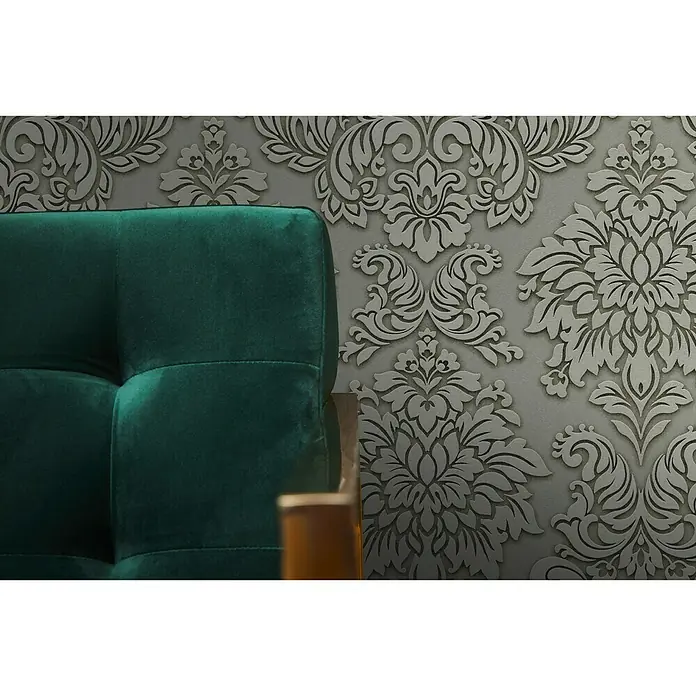 A.S. Création Metropolitan Stories Vliestapete Damask - Grau/Grün, Ornament, 10,05 x 0,53 m - Detailaufnahme eines smaragdgrünen Samtsofas vor einer eleganten grauen Tapete mit floralem Muster, das einen Hauch von Luxus und Komfort vermittelt.