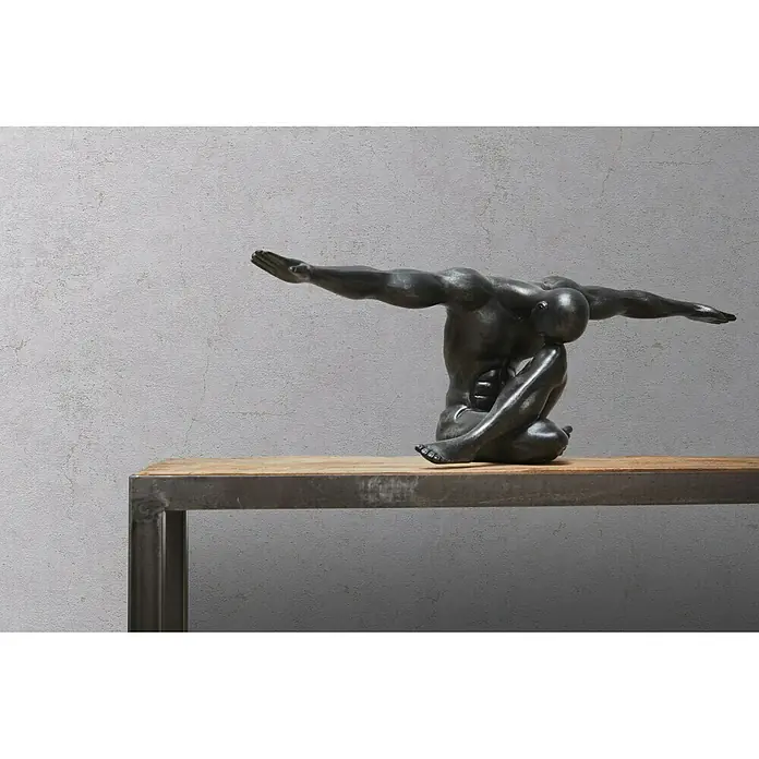 A.S. Création Metropolitan Stories Vliestapete Betonoptik - Taubengrau, Betonoptik, 10,05 x 0,53 m - Bronzefarbene Skulptur eines sitzenden Mannes mit ausgestreckten Armen auf einem Holztisch vor grauer Wand.