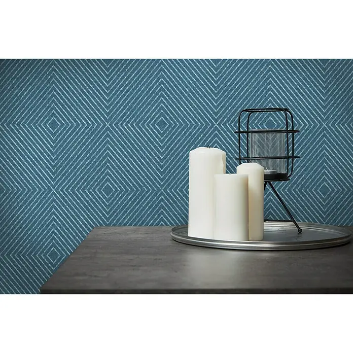 A.S. Création Metropolitan Stories Vliestapete Kachel - Blau, Grafisch, 10,05 x 0,53 m - Nahaufnahme von drei weißen Kerzen und einem schwarzen Kerzenhalter auf einem silbernen Tablett vor einer blauen geometrischen Tapete.