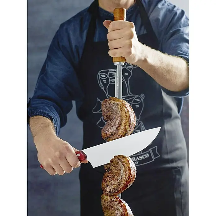 Tramontina Grillspieß - L x B: 75 x 1,7 cm, Edelstahl - Nahaufnahme einer Person, die Churrasco-Fleisch mit einem scharfen Messer von einem Spieß schneidet, während sie eine Grillschürze trägt.