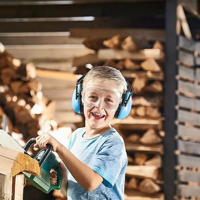 Wolfcraft Gafas de seguridad Kids - Apto para: Niños - Un niño lleva gafas y protectores auditivos, usando felizmente una motosierra de juguete. El equipo es azul y transparente, el juguete verde.