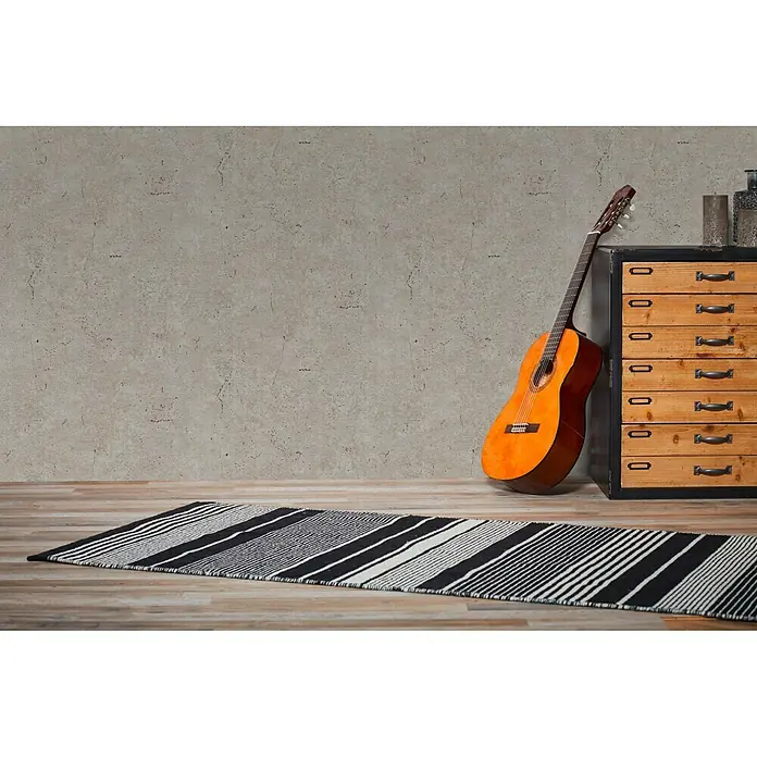 A.S. Création Metropolitan Stories Vliestapete Betonoptik - Taupe, Betonoptik, 10,05 x 0,53 m - Ein gestreifter Teppich in Schwarz und Weiß liegt auf einem Holzboden, mit einer Gitarre und einer Kommode im Hintergrund.