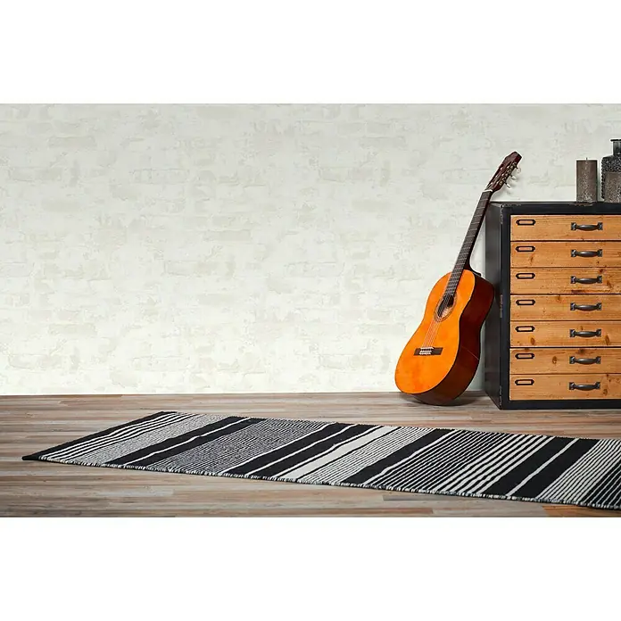 A.S. Création Metropolitan Stories Vliestapete Mauerwerk - Altweiß, Steinoptik, 10,05 x 0,53 m - Gestreifter Teppich in Schwarz und Weiß im Raum mit Gitarre und Kommode. Der Teppich liegt auf dem Holzboden vor einer Wand.