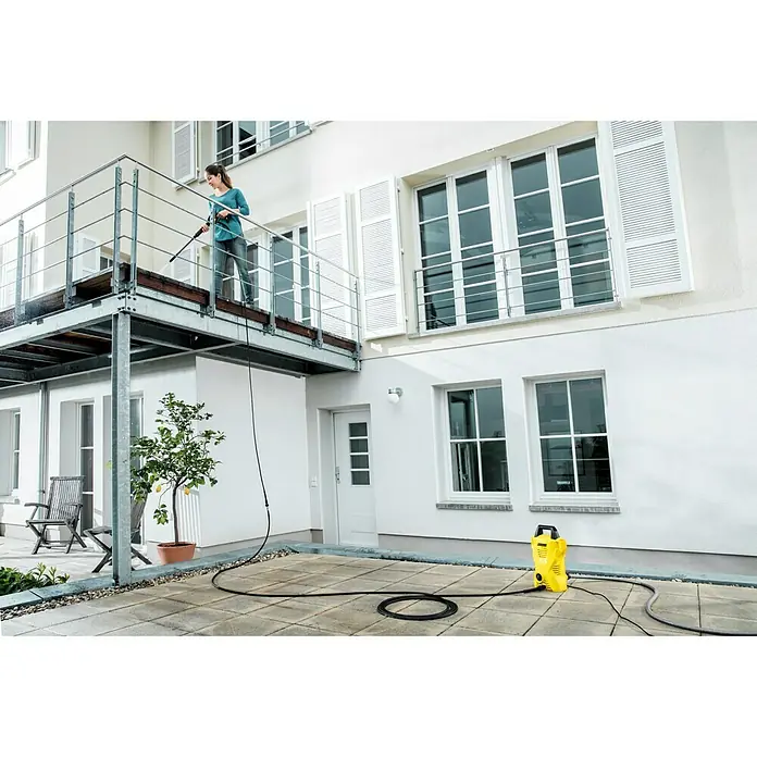 Kärcher Extensión de manguera de alta presión XH 10 - Largo: 10 m, Específico para: Hidrolimpiadora de alta presión Kärcher K2 - K7 vor hasta 2010 - Mujer utiliza una hidrolimpiadora Kärcher en un balcón para limpiar superficies. El dispositivo amarillo está abajo en la terraza.