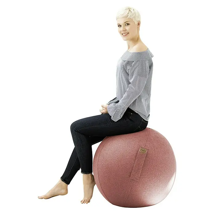 Sitting Ball Gymnastikball Felt - Lachs, Durchmesser: 65 cm, 100 % Polyester - Frau sitzt auf einem Sitzball in Altrosa. Der Ball hat einen Griff und ein kleines Etikett. Sie trägt Jeans und eine gestreifte Bluse.