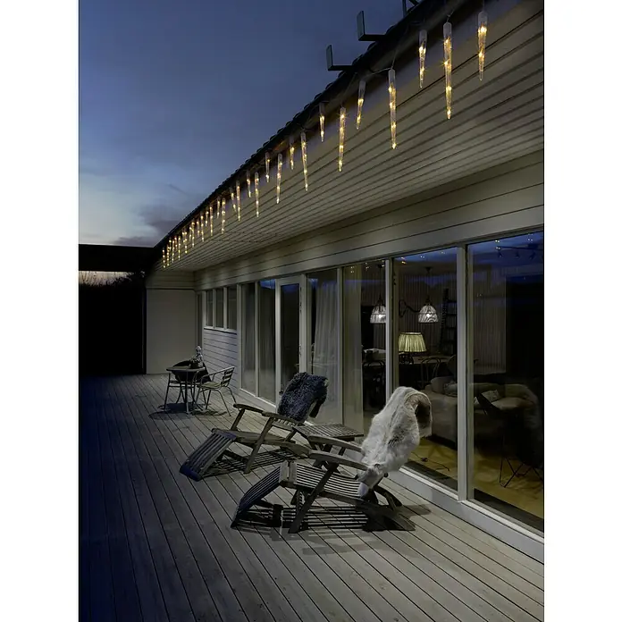 Konstsmide LED-Lichtervorhang Eiszapfen - Außen, 13,75 m, Bernstein, 24 Stk., Netzbetrieben - Beleuchtete Eiszapfen-Lichterketten hängen an einem Haus mit Terrasse und Stühlen und sorgen für eine festliche Atmosphäre.