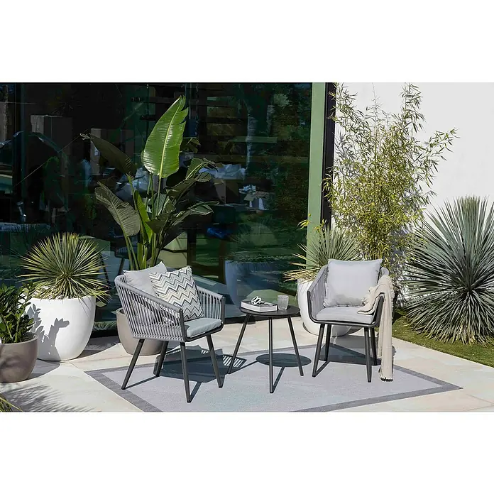 Cojín Zigzag - 45 x 45 cm, Textil, Gris, Cuadrado - Un primer plano de dos sillones grises con una mesa sobre una alfombra en un ambiente exterior rodeado de exuberante vegetación. Los muebles se ven modernos y acogedores.