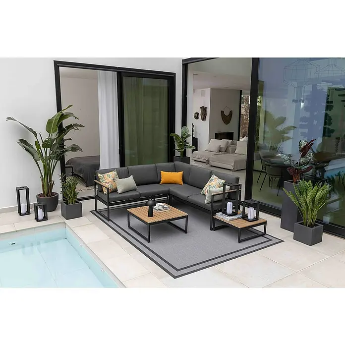 Sunfun Conjunto de muebles Eliane - 5 pzs., Aluminio/Polywood, Antracita - Un elegante conjunto de muebles de exterior con un sofá seccional gris y mesas de madera sobre una alfombra gris junto a una piscina, rodeado de vegetación.