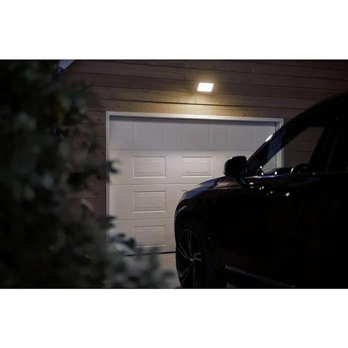 Philips Hue LED-Strahler White & Color Ambiance - 15 W, L x B x H: 22 x 16 x 15,3 cm, Schwarz, RGBW - Ein Garagentor aus Paneelen mit einer Leuchte darüber und einem Teil eines Autos im Bild. Das Tor ist weiß und geschlossen.