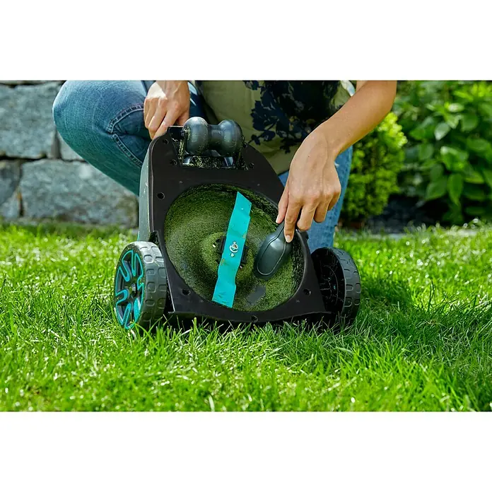 Gardena Ersatzmesser für Rasenmäher - Gardena Akku-Rasenmäher HandyMower Li-18/22 - Eine Frau reinigt einen Rasenmäher mit blauer Klinge. Der Rasenmäher steht auf einem grünen Rasen.