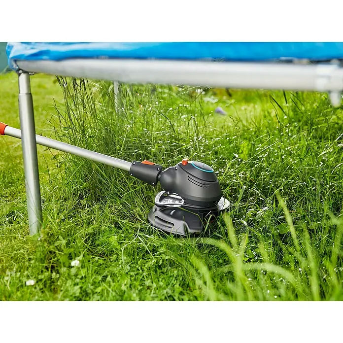 Gardena Elektro-Rasentrimmer ComfortCut 550/28 - 550 W, Schnittbreite Faden: 28 cm - Nahaufnahme eines Gardena Akku-Rasentrimmers im Einsatz unter einem Trampolin, der das Gras präzise schneidet.\n