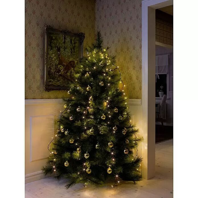 Konstsmide LED-Lichterkette Baummantel - Innen, Lichtfarbe: Bernstein, Anzahl Leuchtmittel: 250 Stk., Netzbetrieben - Weihnachtsbaum geschmückt mit warmweißen Lichtern und goldenen Kugeln, steht in einem gemütlichen Raum.
