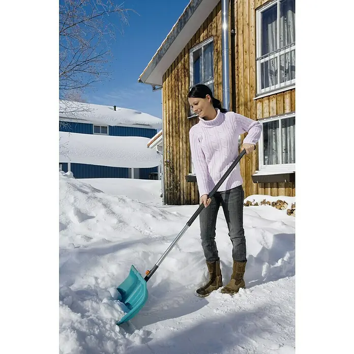 Gardena Combisystem Aluminiumstiel Ergoline 130 - Schwarz/Silber, Material Griff: Kunststoff - Eine Frau schaufelt Schnee mit einer blauen Schneeschaufel vor einem Holzhaus, umgeben von verschneiten Landschaften.