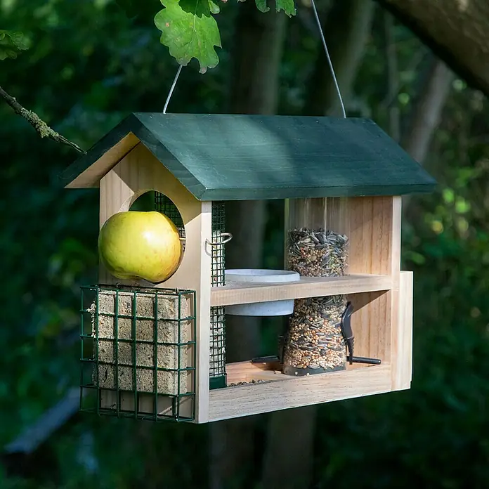 Dobar Futterstation 7-in-1 - 39 x 19 x 29 cm, Natur/Grün - Nahaufnahme eines hölzernen Vogelhauses mit grünem Dach, Apfel, Meisenknödel und Vogelfutter, das an einem Baum hängt.