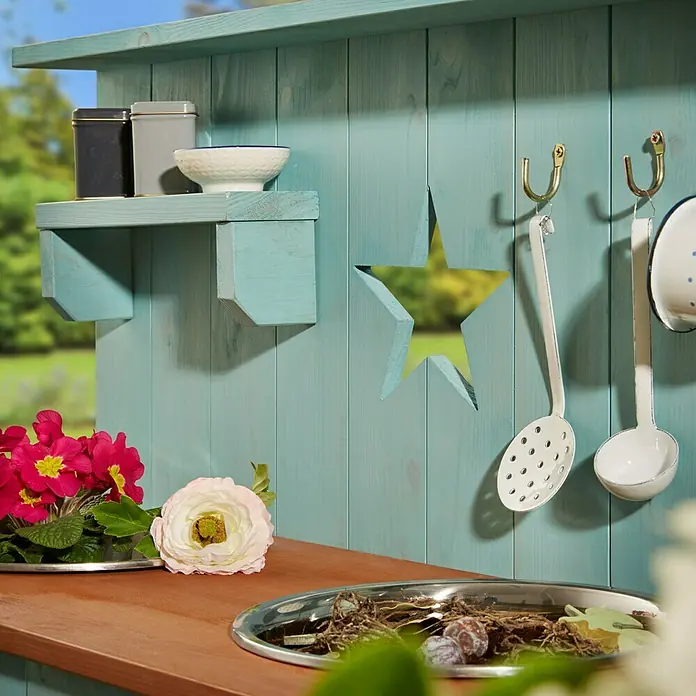 Muddy Buddy Matschküche Adventurer Star - 80 x 40 x 90 cm, Holz, Petrolblau - Detailaufnahme eines bunten Gartenhauses mit Holzregal, Sternaussparung und Küchenutensilien an Haken. Dekorative Blumen und Schalen auf der Arbeitsfläche.