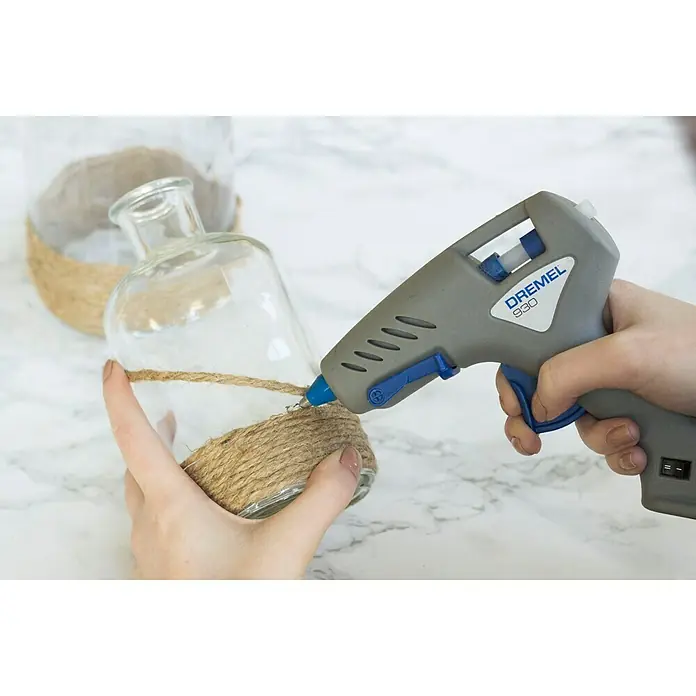 Dremel Pistola termoencoladora Hobby 930-18 - Diámetro cartucho de cola: 7 mm, Tiempo de calentamiento: 5 min - Primer plano de una pistola de pegamento Dremel 930 que se utiliza para pegar cuerda de yute a un jarrón de vidrio.