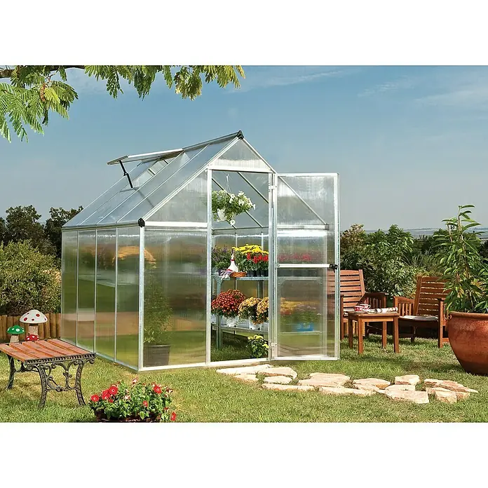 Palram – Canopia Gewächshaus Mythos 6x8 - Außenmaß inkl. Dachüberstand (B x T): 185 x 247 cm, 4 mm, Silber/Transparent - Bild eines Gewächshauses im Garten mit geöffneten Türen, in dem Blumen und Pflanzen auf Regalen wachsen, umgeben von Gartenmöbeln.