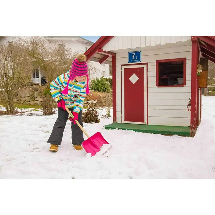 Freund Victoria Kinder-Schneeschieber - Breite: 29 cm, Kurzstiel - Ein Mädchen schippt mit einer pinkfarbenen Schneeschaufel Schnee vor einem kleinen Haus im Garten. Die Schaufel ist ein Blickfang im Winter.