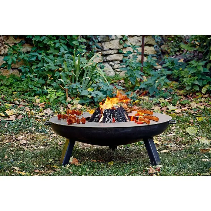 Buschbeck Grillplatte PlanchaStahl, Passend für: Feuerschale mit Ø 80 cm - Eine Feuerschale mit brennendem Holz und Speisen darauf, umgeben von einer grünen Rasenfläche.