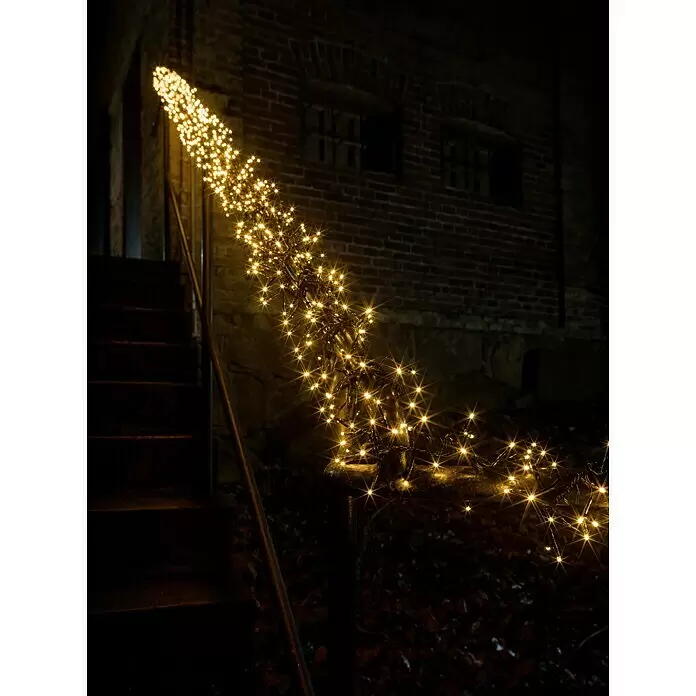 Konstsmide LED-Clusterlichterkette Micro LED Cluster - Außen, Länge: 9,3 m, 576 Stk., Netzbetrieben, Lichtfarbe: Bernstein - Goldene LED-Lichterkette mit Sternenlichteffekt beleuchtet ein Geländer und dunkle Treppen neben einem Backsteingebäude bei Nacht.