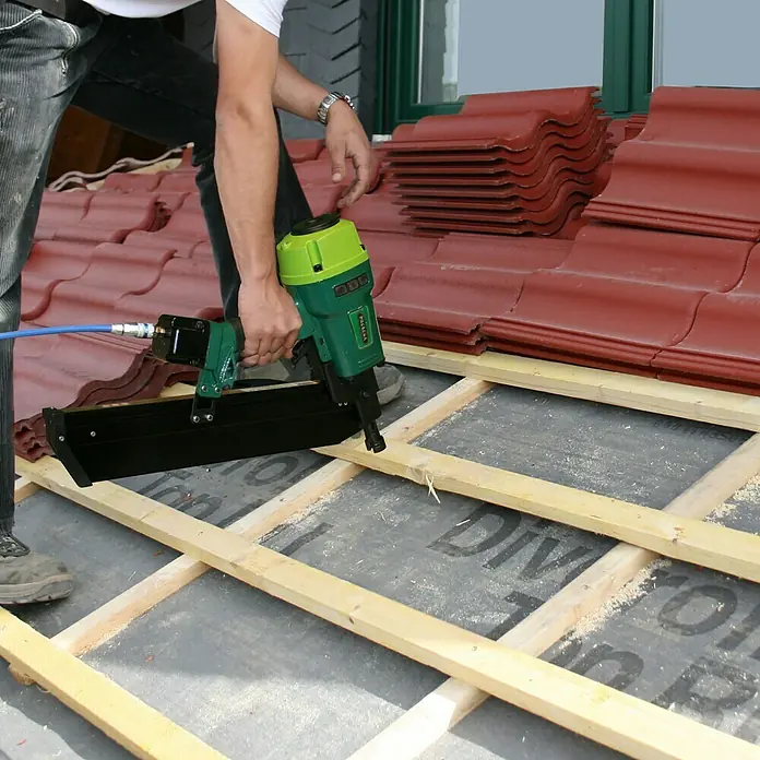Prebena Druckluftnagler 7XR-RK90 - Betriebsdruck: 4 bar - 7 bar, Passend für: Prebena Rundkopf-Streifennägel der Type von RK 50 - 90 mm - Ein Handwerker befestigt Dachlatten mit einem Druckluftnagler auf einem Dach. Der grüne Nagler ist an einen blauen Luftschlauch angeschlossen.