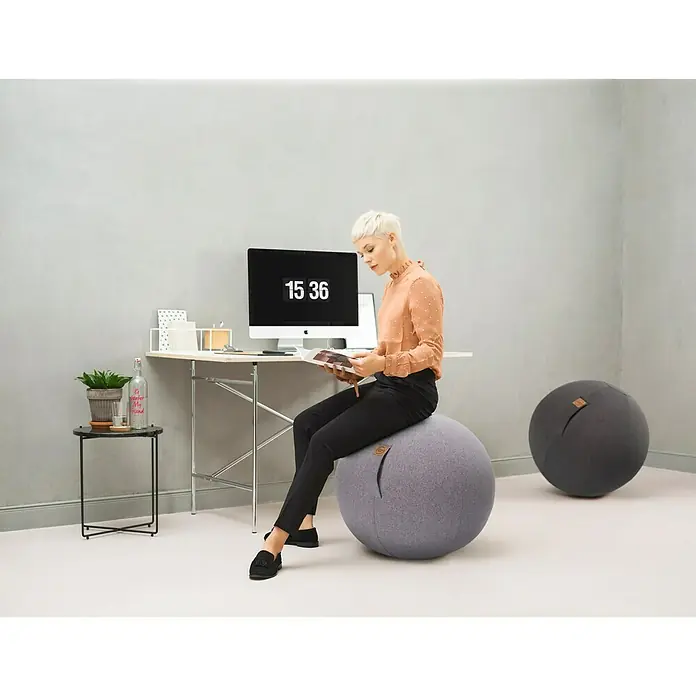 Sitting Ball Gymnastikball Felt - Anthrazit, Durchmesser: 65 cm, 100 % Polyester - Frau sitzt auf einem grauen Gymnastikball mit Stoffbezug vor einem Schreibtisch mit Computer.