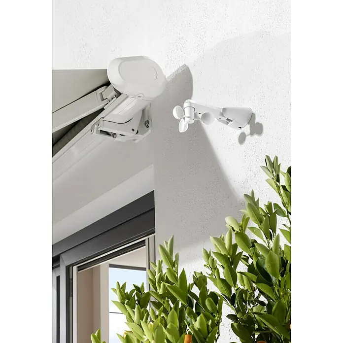 Wind- & Sonnensensor - 130 x 140 x 220 mm, Passend für: Sunfun Vollkassettenmarkisen inkl. LED & Crudo, Sunfun Hülsenmarkisen - Nahaufnahme einer Markise mit einem Windmesser an der Wand. Sonnenschutz für Terrasse und Balkon. Schönes Detail mit Pflanzen.