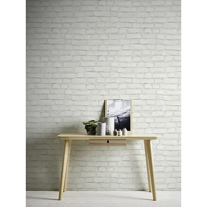 A.S. Création Neue Bude 2.0 ED II Vliestapete Steine - Weiß, Steinoptik, 10,05 x 0,53 m - Ein schmaler, heller Holzschreibtisch vor einer weißen Ziegelwand. Dekorative Elemente auf der Tischplatte wie Pflanzen und ein Bild.