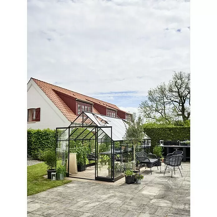 Halls Gewächshaus Qube 816 - Außenmaß inkl. Dachüberstand (B x T): 261 x 508 cm, 3 mm, Schwarz - Schwarz gerahmtes Gewächshaus mit klaren Scheiben, Pflanzen im Inneren, in einem Garten mit Sitzgelegenheiten auf einer Terrasse.