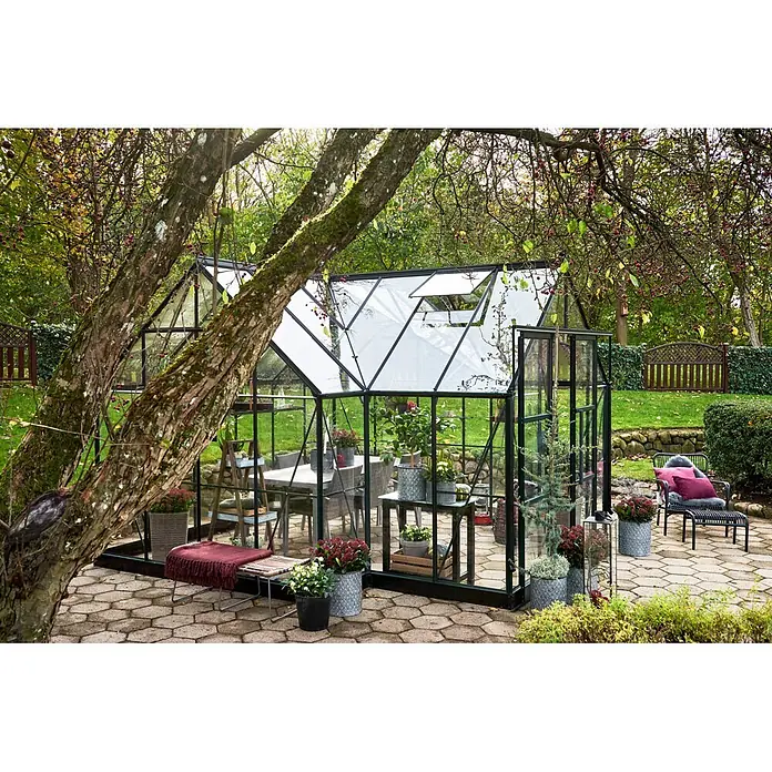 Halls Gewächshaus Garden Room - Außenmaß inkl. Dachüberstand (B x T): 389 x 390 cm, Farbe Gestell: Grün, Wandstärke: 3 mm - Gewächshaus mit schwarzem Rahmen und Glaspaneelen, innen eingerichtet, in einem üppigen Garten.