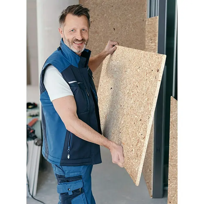 elka ESB-Platte Plus - Ein Handwerker hält eine OSB-Platte für den Innenausbau in der Hand, trägt blaue Arbeitskleidung und lächelt in die Kamera.