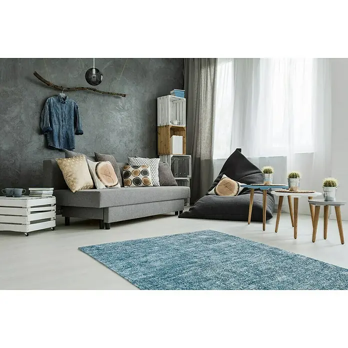 Kayoom Kurzflorteppich Etna - Petrol, 150 x 80 cm, 100 % Mikropolyester - Ein türkisfarbener Teppich liegt in einem modernen Wohnzimmer mit grauer Couch, Sitzsack und Holztischen. Der Teppich bringt Farbe in den Raum.