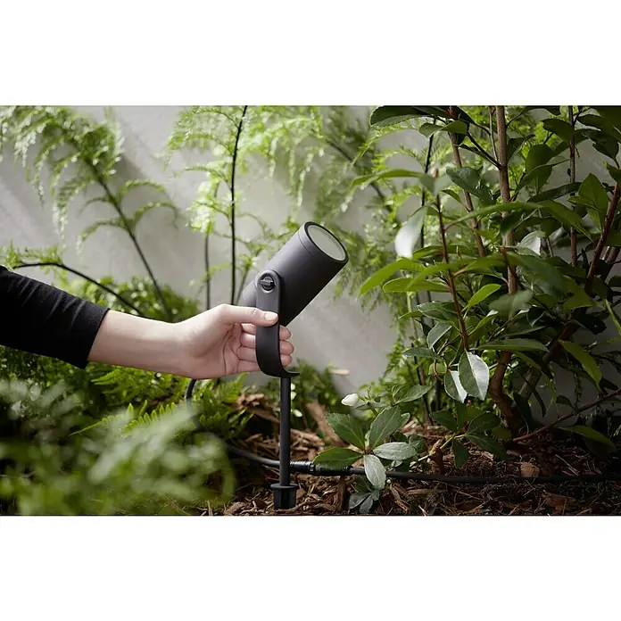 Philips Hue LED-Gartenspot Lily - 8 W, Lichtfarbe: RGB, IP65 - Ein schwarzer LED-Gartenstrahler wird in einem Garten mit üppigem Grün aufgestellt. Der Strahler beleuchtet Pflanzen und Büsche wirkungsvoll.