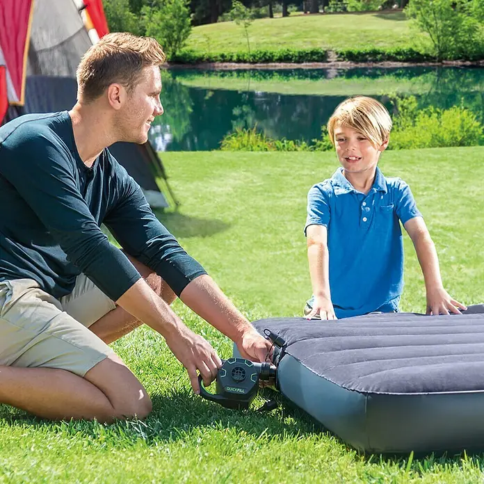 Intex Bomba de aire Quick Fill - 3 boquillas, 230 V - Un padre está inflando un colchón de aire gris con una bomba de aire QuickFill con su hijo delante de una tienda en un prado verde.