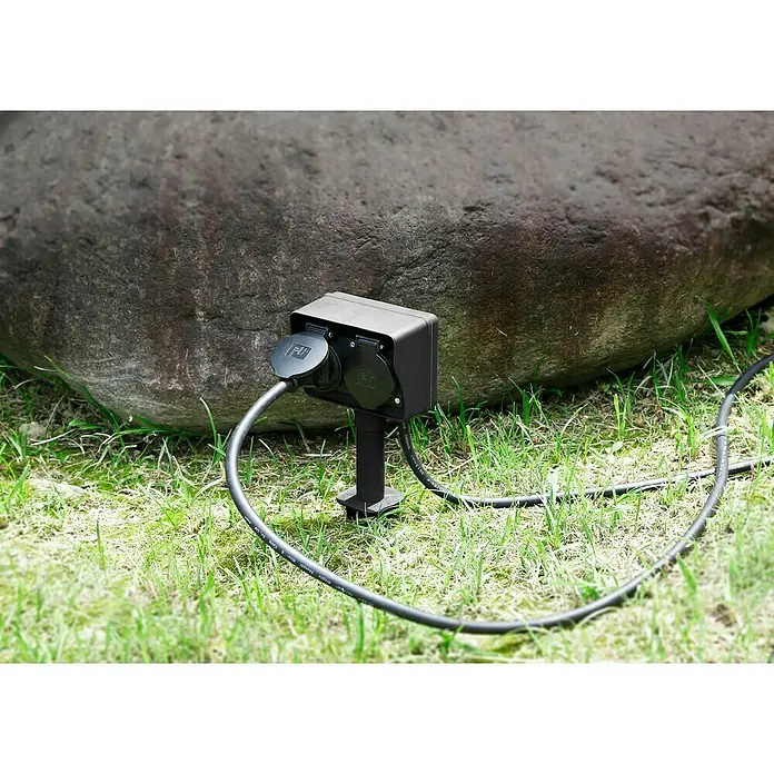 Voltomat Erdspieß-Steckdose - Kabellänge: 1,5 m, 2 Stk., IP44, 2-fach - Outdoor-Steckdose mit zwei Anschlüssen und schwarzem Kabel auf Rasen vor einem großen Stein.