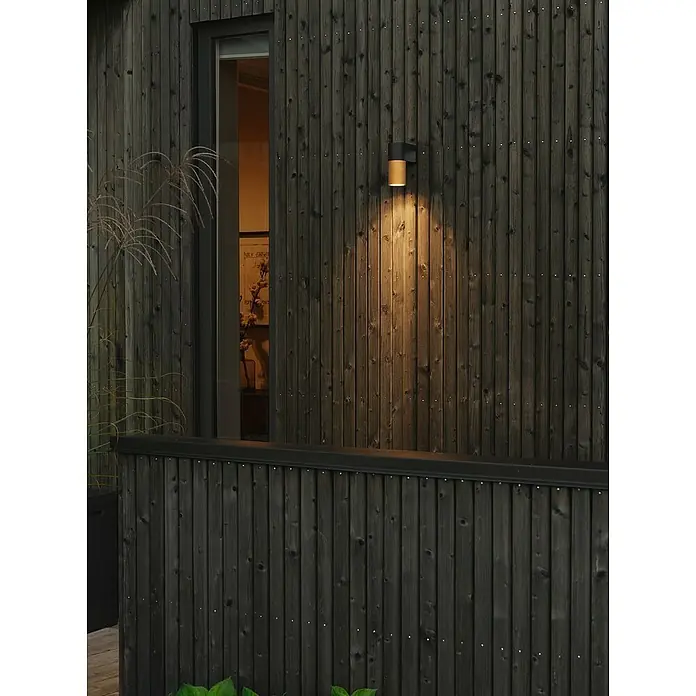 Konstsmide Außenwandleuchte Temi - 7 W, Schwarz/Gold, IP54 - Diese Aufnahme zeigt eine stilvolle, zweifarbige Außenwandleuchte an einer Fassade aus dunklem Holz, die warmes Licht nach unten abstrahlt.