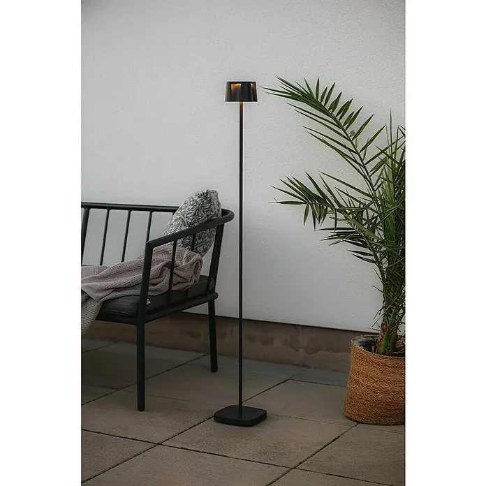 Konstsmide Akku-Stehleuchte Nice - 10 x 10 x 120 cm, Schwarz, Mehrfarbig, 3 W, IP54 - Eine moderne, schwarze Stehlampe mit einem minimalistischen Design, die neben einem Sessel und einer Pflanze auf einer Terrasse steht.