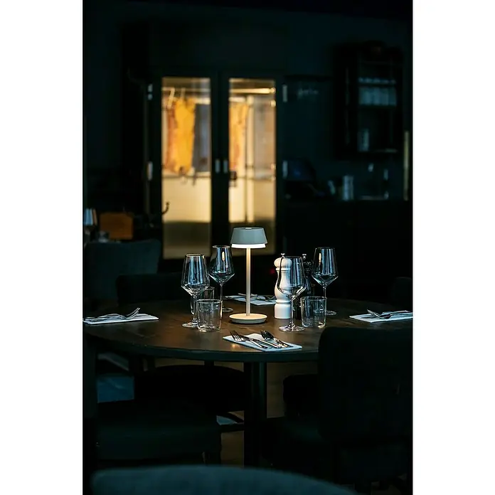 Konstsmide Akku-Tischleuchte Lyon - 2,5 W, L x B x H: 12 x 12 x 30 cm, Sand, RGBW - Ein gedeckter Tisch mit einem modernen, weißen Akku-Lampe, Wein- und Wassergläsern sowie Besteck in einem eleganten Restaurant.