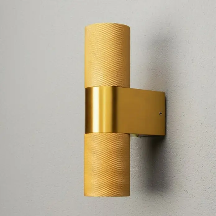 Konstsmide Außenwandleuchte Temi - 14 W, Gold, IP54 - Nahaufnahme einer goldenen Wandleuchte mit zylindrischen Lampenschirmen und einem rechteckigen Sockel an einer grauen Wand.