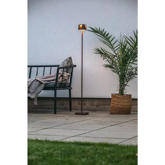 Konstsmide Akku-Stehleuchte Nice - 10 x 10 x 120 cm, Rost, Mehrfarbig, 3 W, IP54 - Eine moderne Stehlampe mit warmem Licht steht neben einer Bank und einer Pflanze auf einer Terrasse. Die Lampe sorgt für eine gemütliche Atmosphäre im Außenbereich.